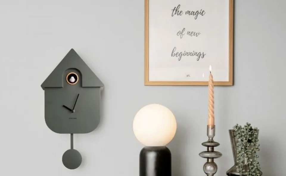 Nowoczesna dekoracja z zegarem: Zegar ścienny w kształcie domku kukułki, wiszący obok dekoracyjnej świecy i lampy, na tle jasnej ściany z napisem „the magic of new beginnings”