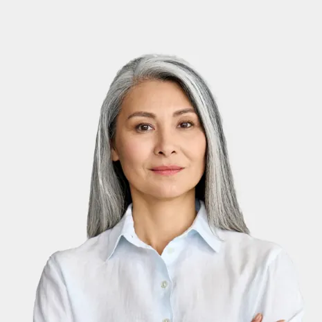 Portret kobiety: Kobieta o długich, siwych włosach, ubrana w białą koszulę, z założonymi rękami, patrząca prosto w obiektyw na neutralnym tle.